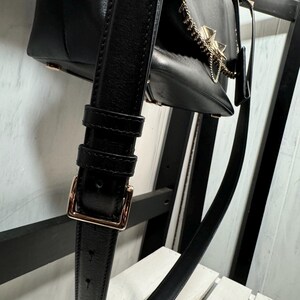 May include: Black leather handbag with a gold-tone chain and accents. The bag features a long, adjustable strap with a gold buckle. The bag is hanging on a black wooden structure.