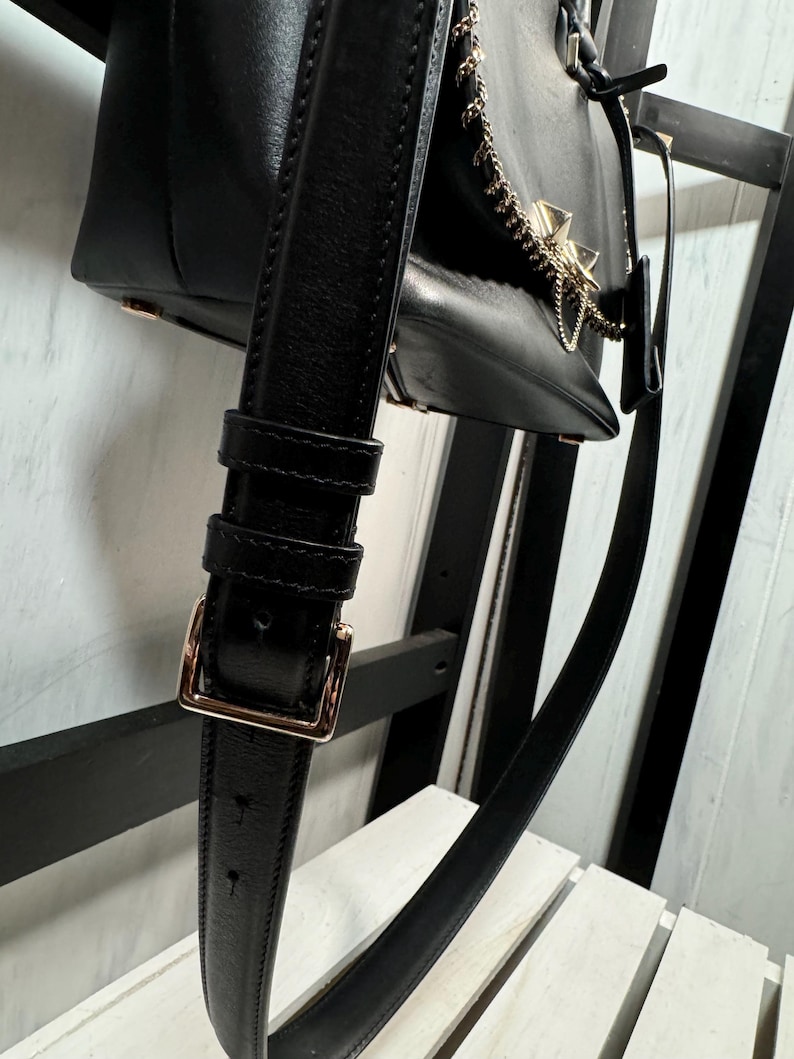 May include: Black leather handbag with a gold-tone chain and accents. The bag features a long, adjustable strap with a gold buckle. The bag is hanging on a black wooden structure.