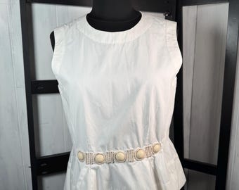 Peut inclure: Haut sans manches blanc avec un col rond et une ceinture décorative ornée de gros boutons ronds de couleur crème. Le haut a un ourlet de style péplum et est présenté sur un mannequin noir.