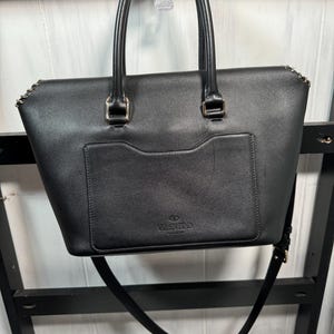 May include: Black leather tote bag with top handles and a detachable shoulder strap. The bag features a front pocket and gold-tone hardware accents. The brand name "Valentino" is embossed on the front.