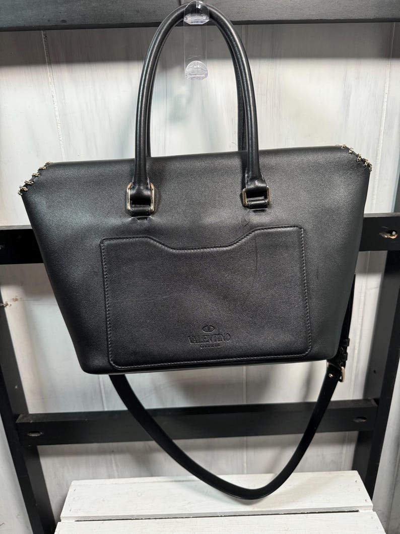 May include: Black leather tote bag with top handles and a detachable shoulder strap. The bag features a front pocket and gold-tone hardware accents. The brand name "Valentino" is embossed on the front.