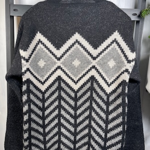Puede incluir: Un suéter gris oscuro con un patrón geométrico en blanco, gris claro y gris oscuro. El patrón presenta formas de diamante en la parte superior y un diseño de cheurón debajo. El suéter está colgado en una percha negra.