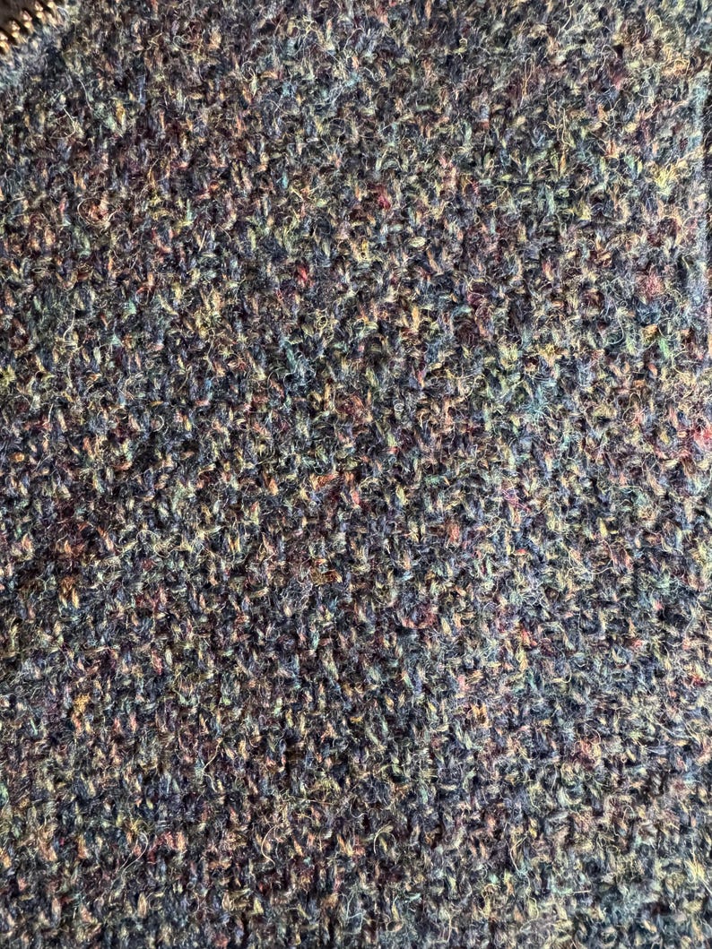 May include: Close-up of a textured fabric with a mix of colors. The fabric appears to be woven with a variety of threads, including shades of blue, green, purple, and hints of red and yellow. The texture is dense and slightly uneven.
