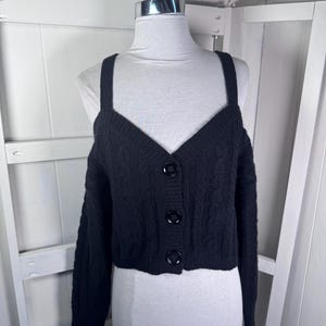 May include: A black, open-shoulder cardigan with long sleeves and a V-neckline. The cropped sweater features a cable knit design and three large, round buttons down the front. The garment is displayed on a mannequin.