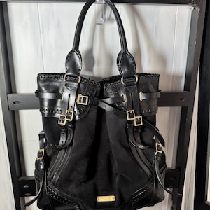 Puede incluir: Bolso tote negro con correas de cuero, hebillas doradas y un centro de ante. El bolso tiene dos asas superiores y una placa rectangular dorada en la parte delantera. El bolso cuelga sobre un fondo blanco y negro.