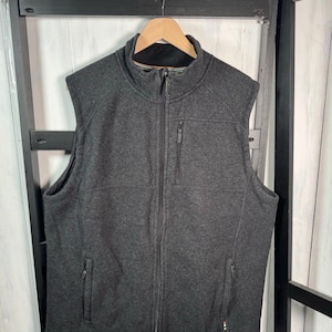 May include: A charcoal gray fleece vest with a full-length zipper and a stand-up collar. The vest has two side pockets and a chest pocket, and is hanging on a wooden hanger.