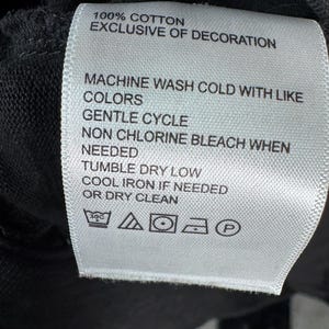 May include: A white fabric tag with black text, attached to a dark garment. The tag reads "100% COTTON EXCLUSIVE OF DECORATION" and provides washing instructions: machine wash cold, gentle cycle, non-chlorine bleach, tumble dry low, cool iron, or dry clean.