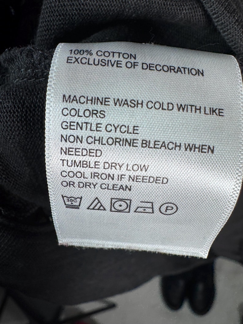 May include: A white fabric tag with black text, attached to a dark garment. The tag reads "100% COTTON EXCLUSIVE OF DECORATION" and provides washing instructions: machine wash cold, gentle cycle, non-chlorine bleach, tumble dry low, cool iron, or dry clean.
