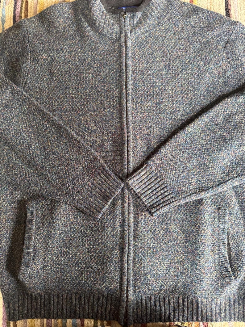 May include: A blue and gray zip-up sweater with a textured knit pattern. The sweater has a high collar, two front pockets, and ribbed cuffs and hem. The zipper runs down the center of the sweater.