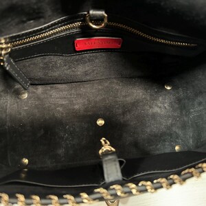 May include: Black leather tote bag with gold-tone chain strap and hardware. The interior features a zippered pocket and a red label with the brand name. The bag has a spacious interior and a classic design.