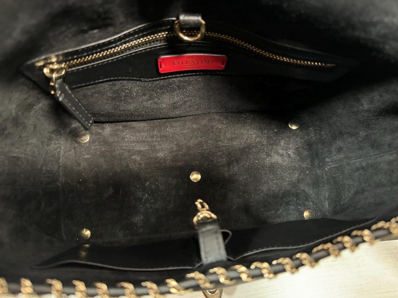 May include: Black leather tote bag with gold-tone chain strap and hardware. The interior features a zippered pocket and a red label with the brand name. The bag has a spacious interior and a classic design.
