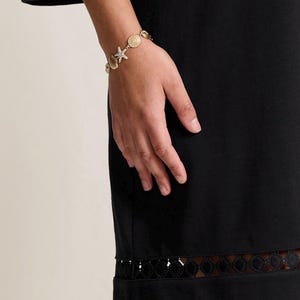 May include: Gold-toned bracelet featuring seashell, starfish, and spiral charms. The bracelet is worn on a person's wrist, with a black dress in the background. The dress has a decorative trim at the hem and sleeve.