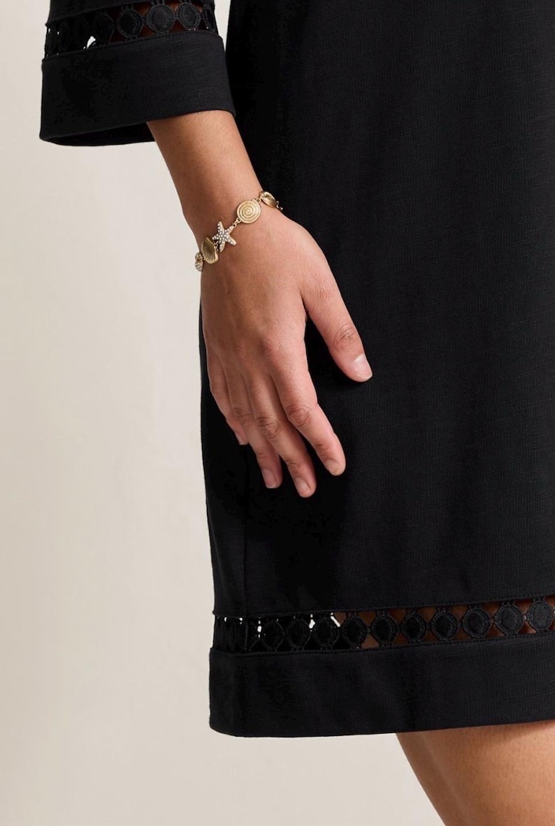 May include: Gold-toned bracelet featuring seashell, starfish, and spiral charms. The bracelet is worn on a person's wrist, with a black dress in the background. The dress has a decorative trim at the hem and sleeve.