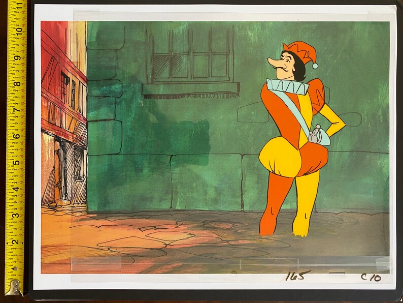 May include: Animation cel of a cartoon jester in a colorful costume. The jester wears a red and yellow hat, a white ruffled collar, and a long nose. The background depicts a green wall and a building with a red roof.