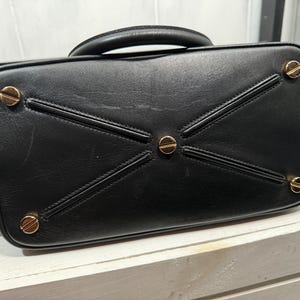 May include: Black leather handbag with a top handle and gold-toned hardware. The bottom of the bag features a geometric design with gold accents. The bag's base has four gold-colored feet.