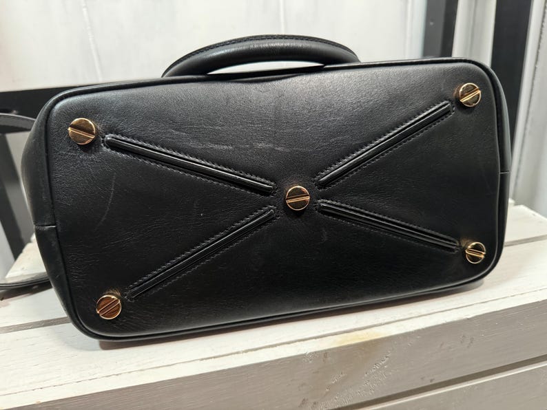 May include: Black leather handbag with a top handle and gold-toned hardware. The bottom of the bag features a geometric design with gold accents. The bag's base has four gold-colored feet.