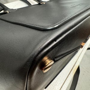 May include: A black leather briefcase with a top handle and gold-colored hardware. The bag has a flap closure and a structured design. The hardware includes decorative studs and a clasp. The briefcase is likely designed for carrying documents and other professional items.