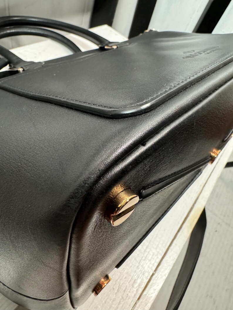 May include: A black leather briefcase with a top handle and gold-colored hardware. The bag has a flap closure and a structured design. The hardware includes decorative studs and a clasp. The briefcase is likely designed for carrying documents and other professional items.