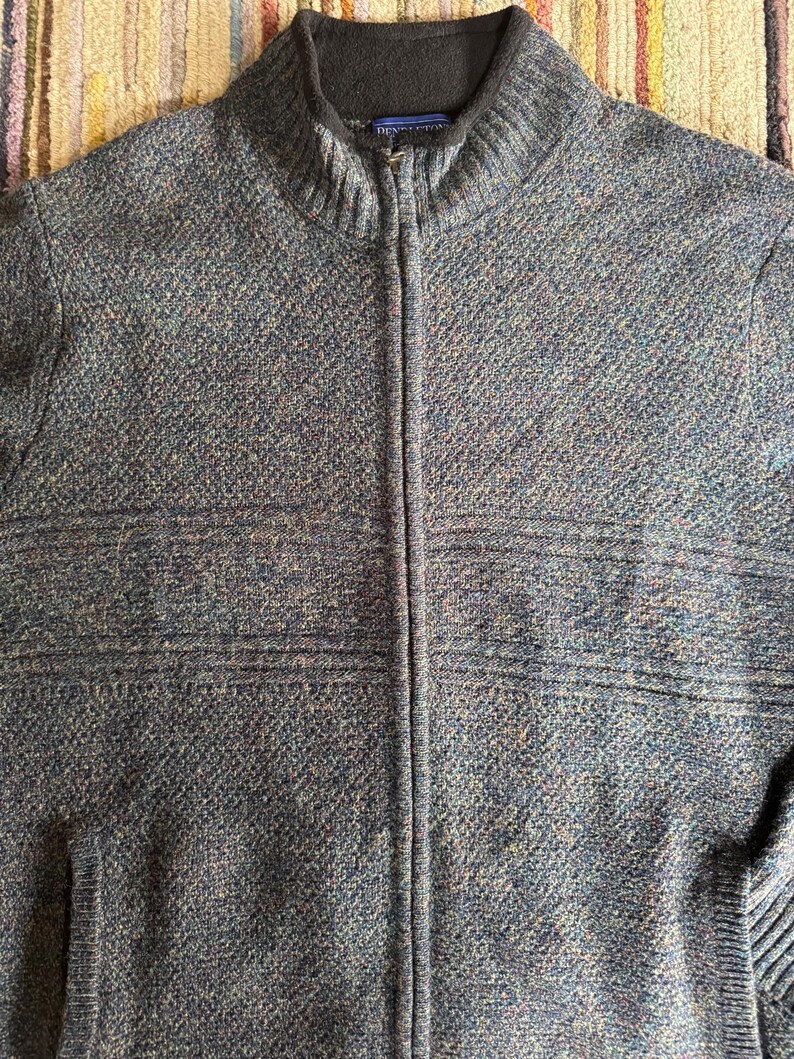 May include: A blue and gray zip-up sweater with a high collar. The sweater has a textured knit pattern and a full-length zipper. The brand name "Pendleton" is visible on the collar. The sweater has two front pockets.