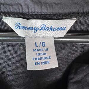 May include: Close-up of a black garment tag with the brand name "Tommy Bahama" in blue script. The tag also indicates the size "L/G" and "Made in India / Fabriqu&eacute; en Inde". The garment is black.