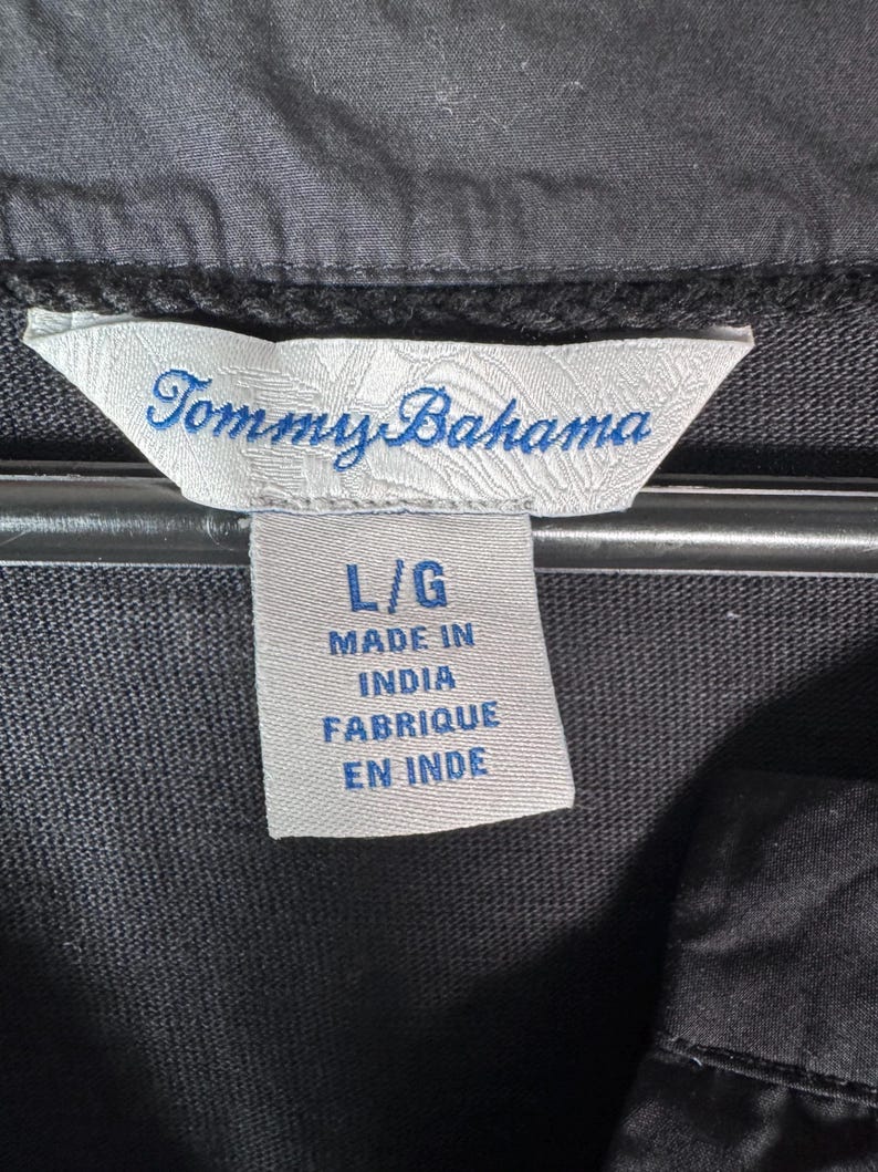May include: Close-up of a black garment tag with the brand name "Tommy Bahama" in blue script. The tag also indicates the size "L/G" and "Made in India / Fabriqu&eacute; en Inde". The garment is black.