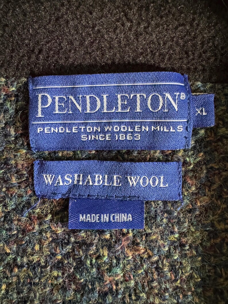 May include: Close-up of a blue label with the word "PENDLETON" in white, and the text "PENDLETON WOOLEN MILLS SINCE 1863". Below is a smaller label that reads "WASHABLE WOOL" and "MADE IN CHINA". The background is a textured fabric.