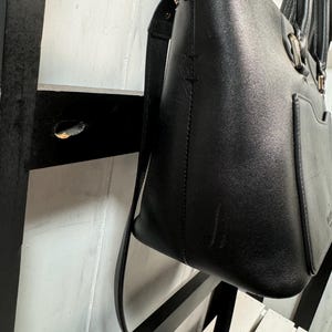 May include: Black leather tote bag with a shoulder strap and chain detail on the top edge. The bag has a rectangular shape with a pocket on the front and a handle. The bag is hanging against a white and black striped background.