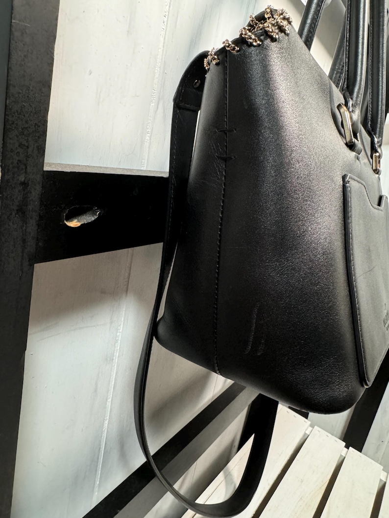 May include: Black leather tote bag with a shoulder strap and chain detail on the top edge. The bag has a rectangular shape with a pocket on the front and a handle. The bag is hanging against a white and black striped background.