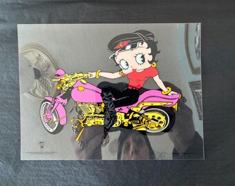 May include: A pop art style image of Betty Boop riding a pink motorcycle with gold accents. Betty Boop is wearing a red top, black pants, and a black hat. The motorcycle has a pink frame and gold engine components.
