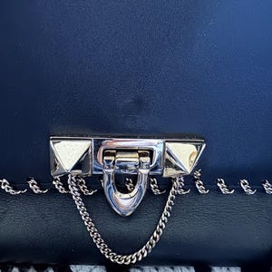 May include: Close-up of a navy blue handbag with a silver clasp and chain detail. The clasp has pyramid-shaped studs on each side. The bag is made of a textured material.