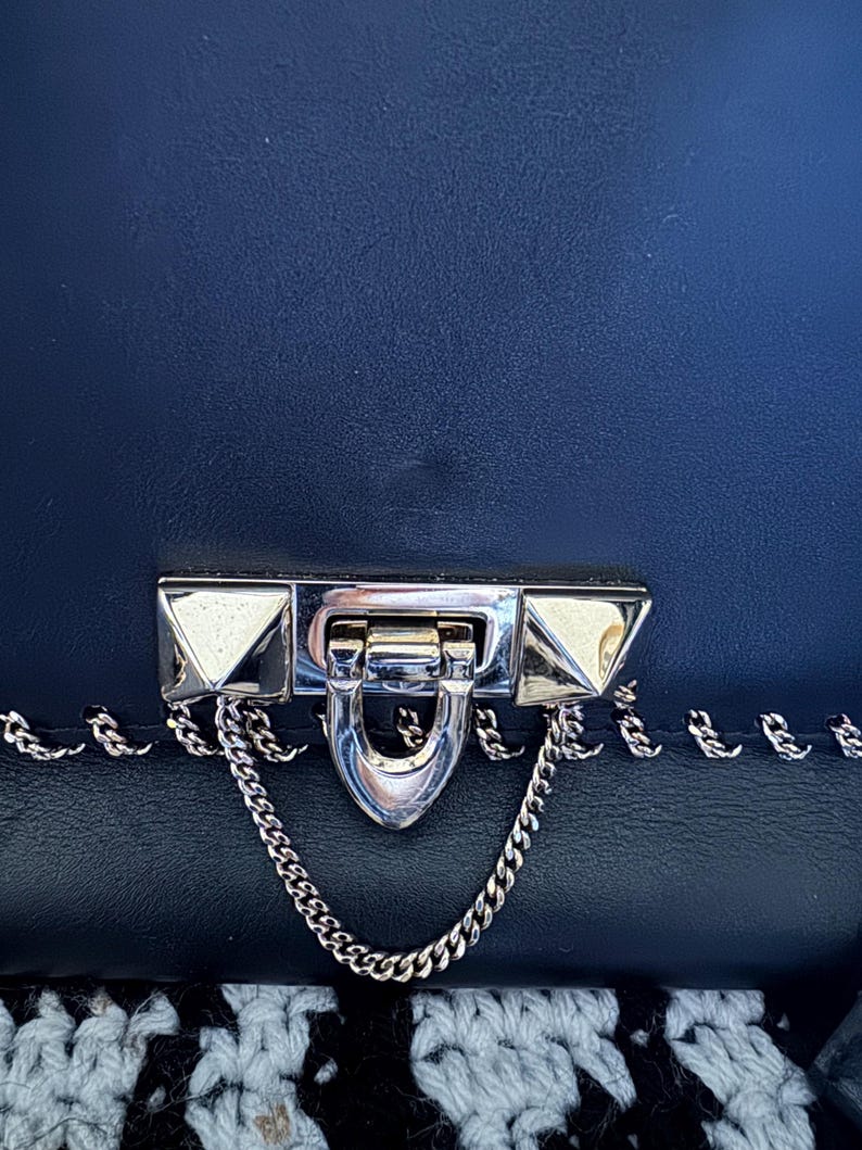 May include: Close-up of a navy blue handbag with a silver clasp and chain detail. The clasp has pyramid-shaped studs on each side. The bag is made of a textured material.