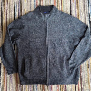 May include: A gray zip-up sweater with a high collar and two front pockets. The sweater has a textured knit pattern and horizontal stripe detailing across the chest. The zipper runs down the center of the sweater.