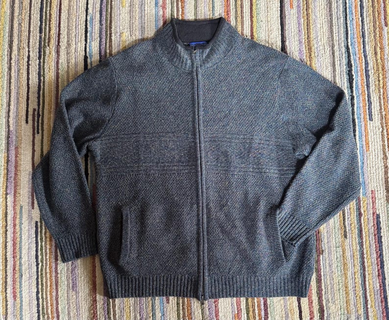 May include: A gray zip-up sweater with a high collar and two front pockets. The sweater has a textured knit pattern and horizontal stripe detailing across the chest. The zipper runs down the center of the sweater.