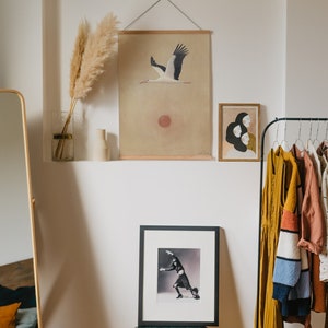 May include: A room interior featuring a wall niche with decorative items. A framed print of a stork in flight hangs above a framed black and white photograph. A gold-framed mirror and a clothing rack with various garments are also visible.