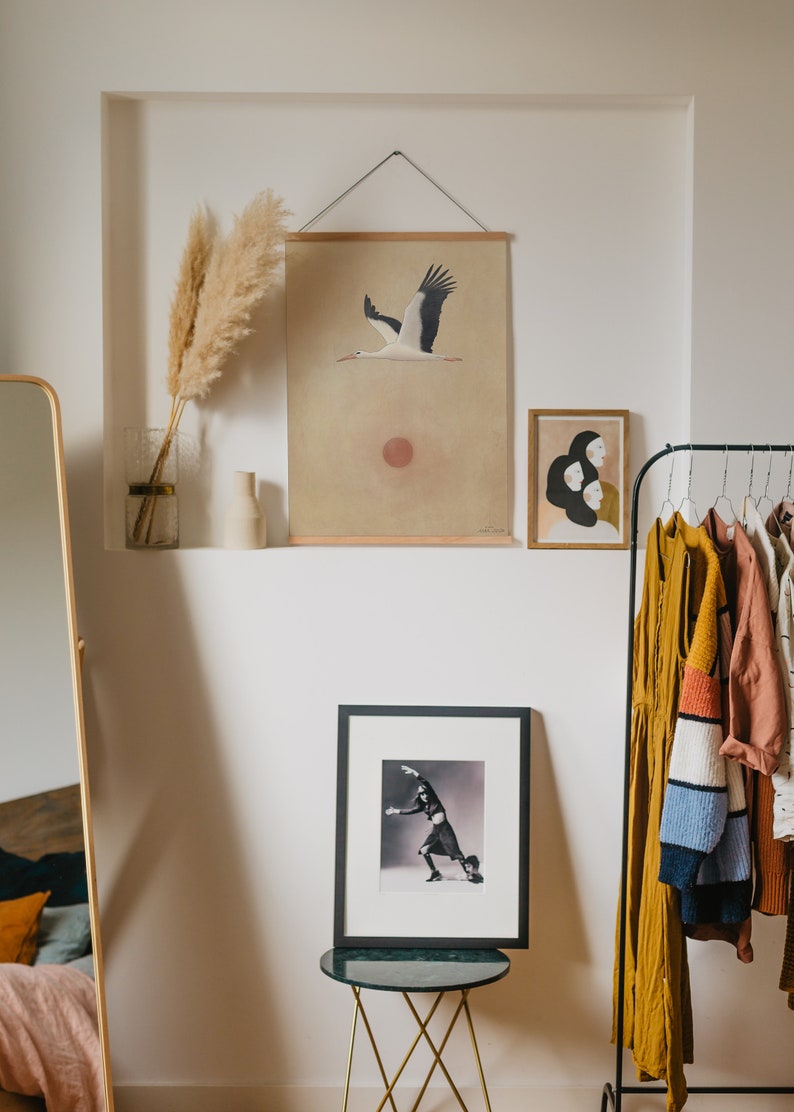 May include: A room interior featuring a wall niche with decorative items. A framed print of a stork in flight hangs above a framed black and white photograph. A gold-framed mirror and a clothing rack with various garments are also visible.