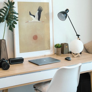 May include: A modern home office setup featuring a wooden desk with a framed art print of a flying stork. The desk also holds a laptop, lamp, speaker, camera, and a white chair.