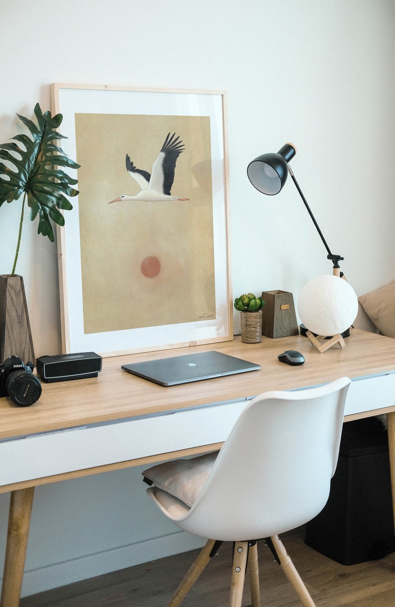 May include: A modern home office setup featuring a wooden desk with a framed art print of a flying stork. The desk also holds a laptop, lamp, speaker, camera, and a white chair.