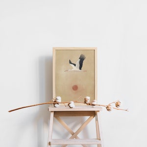 May include: A framed art print featuring a stork in flight against a beige background with a red sun. The artwork is displayed on a wooden step stool, with a branch of cotton blossoms in the foreground. The print has a natural, minimalist aesthetic.