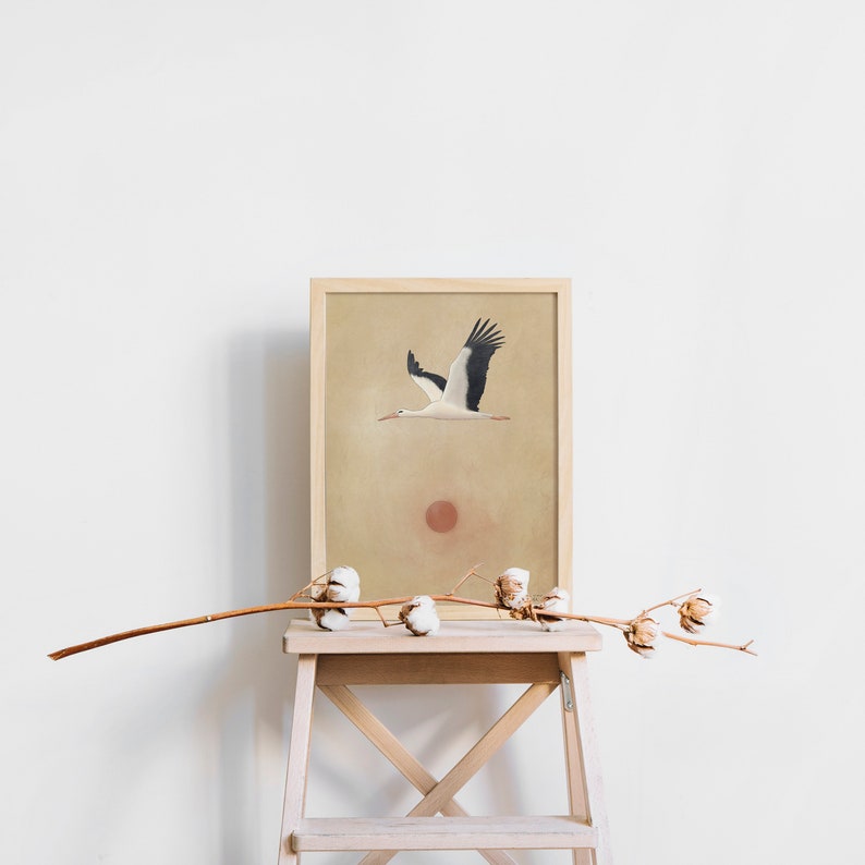 May include: A framed art print featuring a stork in flight against a beige background with a red sun. The artwork is displayed on a wooden step stool, with a branch of cotton blossoms in the foreground. The print has a natural, minimalist aesthetic.