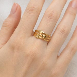 Puede incluir: Un anillo de tono dorado con un dise&ntilde;o de banda ondulada y org&aacute;nica. El anillo est&aacute; grabado con la letra S y un s&iacute;mbolo del zodiaco Virgo. La superficie met&aacute;lica tiene un acabado pulido y la banda consta de dos l&iacute;neas fluidas entrelazadas.