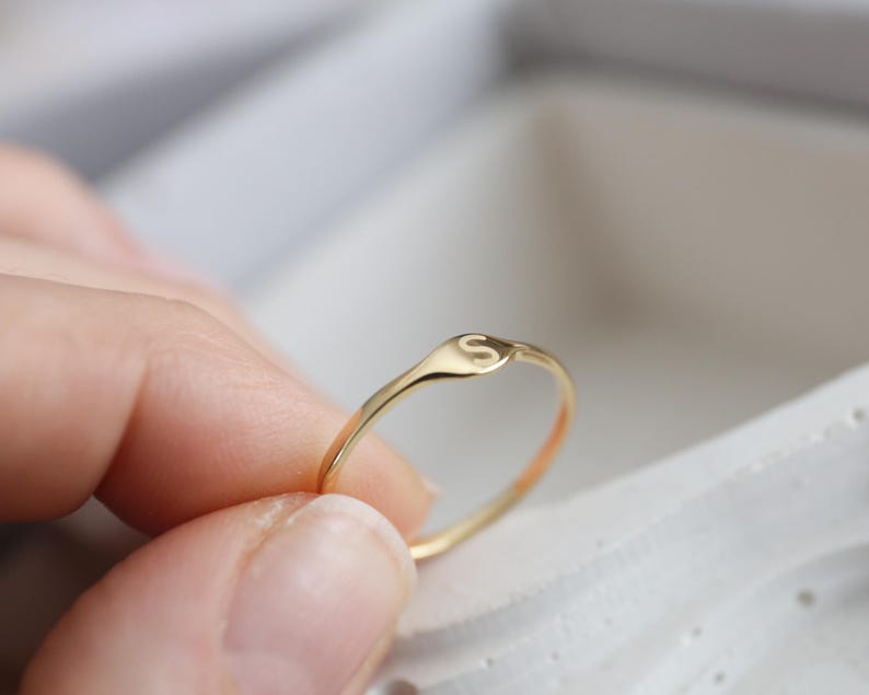 Puede incluir: Un anillo fino de oro con una peque&ntilde;a cara circular plana grabada con la letra may&uacute;scula S. La banda es lisa y pulida, mostrando un dise&ntilde;o sencillo con un perfil delicado y un acabado met&aacute;lico reflectante.