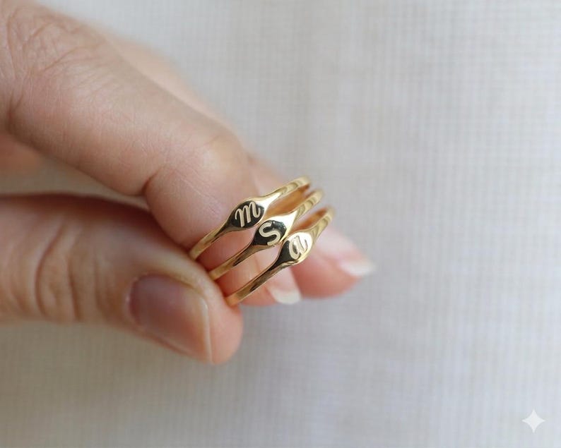 Puede incluir: Tres anillos de tono dorado con bandas delgadas y caras peque&ntilde;as, planas y ovaladas. Cada cara del anillo est&aacute; grabada con una sola letra min&uacute;scula en cursiva, espec&iacute;ficamente las letras m, s y a, dispuestas en formaci&oacute;n apilada.