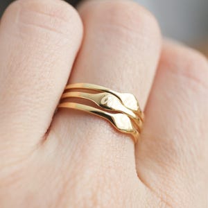 Puede incluir: Un conjunto de tres anillos dorados apilados, cada uno con un acabado liso y pulido y una peque&ntilde;a placa rectangular plana centrada. El anillo superior est&aacute; grabado con una letra cursiva, mientras que los otros dos anillos permanecen lisos.