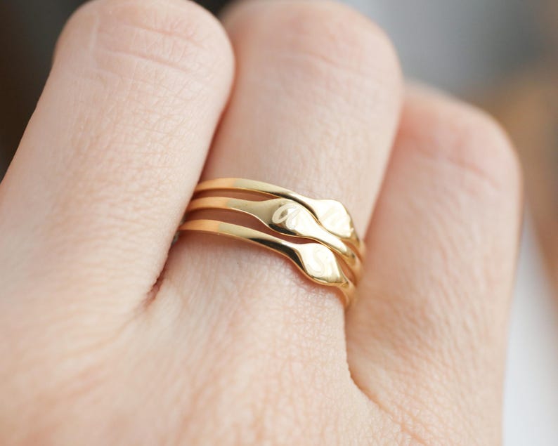 Puede incluir: Un conjunto de tres anillos dorados apilados, cada uno con un acabado liso y pulido y una peque&ntilde;a placa rectangular plana centrada. El anillo superior est&aacute; grabado con una letra cursiva, mientras que los otros dos anillos permanecen lisos.