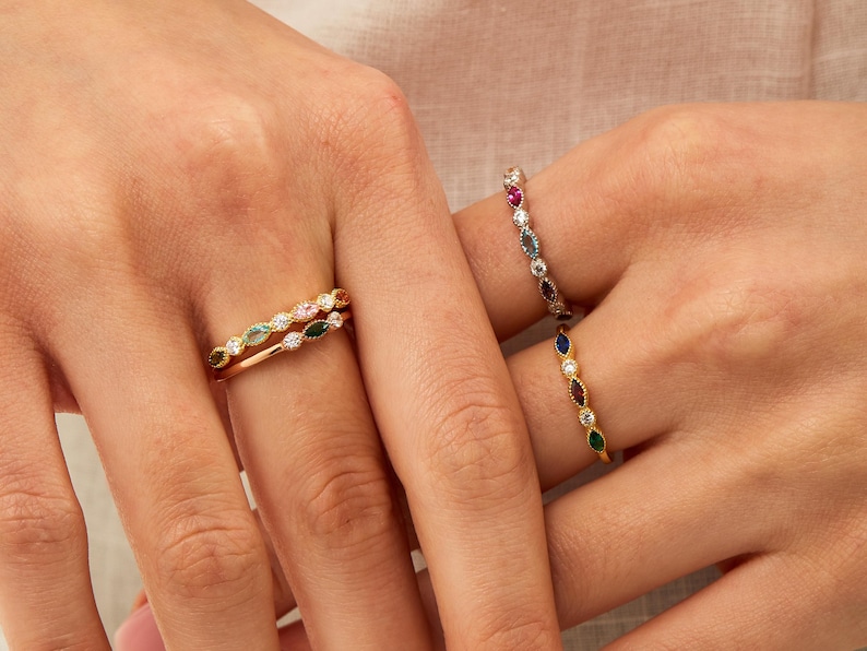 Peut inclure: Trois bagues en or avec des pierres pr&eacute;cieuses color&eacute;es. Les bagues sont empil&eacute;es sur un doigt. La bague du haut a des pierres pr&eacute;cieuses vertes, roses et blanches. La bague du milieu a des pierres pr&eacute;cieuses vertes. La bague du bas a des pierres pr&eacute;cieuses bleues et vertes.