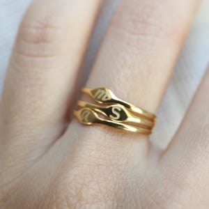Puede incluir: Un conjunto de tres anillos finos de tono dorado apilados. Cada anillo presenta una peque&ntilde;a cara plana de forma ovalada grabada con una letra min&uacute;scula. Las letras visibles en los anillos son m, s y a, en una fuente sencilla y clara.