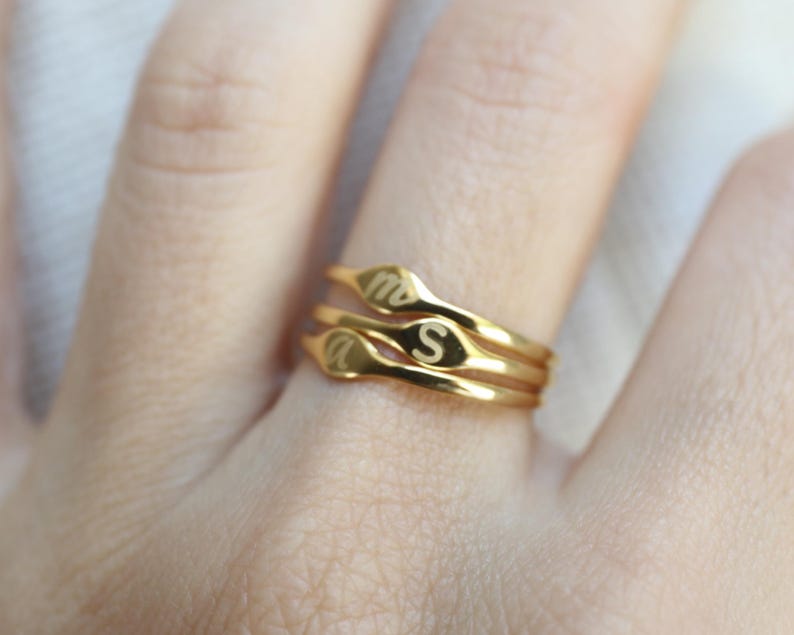 Puede incluir: Un conjunto de tres anillos finos de tono dorado apilados. Cada anillo presenta una peque&ntilde;a cara plana de forma ovalada grabada con una letra min&uacute;scula. Las letras visibles en los anillos son m, s y a, en una fuente sencilla y clara.
