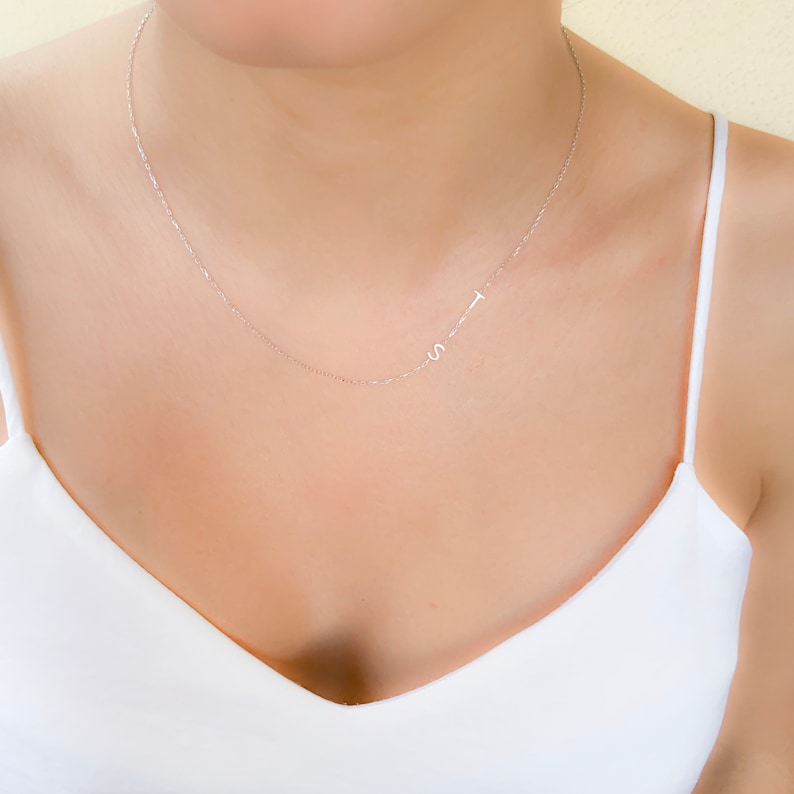 Puede incluir: Un delicado collar de cadena de plata con un peque&ntilde;o dije de cruz y un dije de letra 'S'. El collar lo lleva una persona con una camiseta sin mangas blanca.