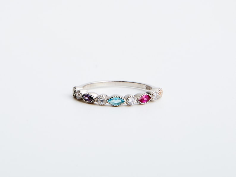 Peut inclure: Une bague en argent avec une rang&eacute;e de pierres pr&eacute;cieuses de forme marquise color&eacute;es. La bague pr&eacute;sente une pierre pr&eacute;cieuse violette, bleue et rouge, avec des pierres rondes claires entre les deux.