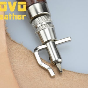 May include: A close-up of a silver leather stitching tool with a wooden handle. The tool is being used to stitch a piece of light brown leather.